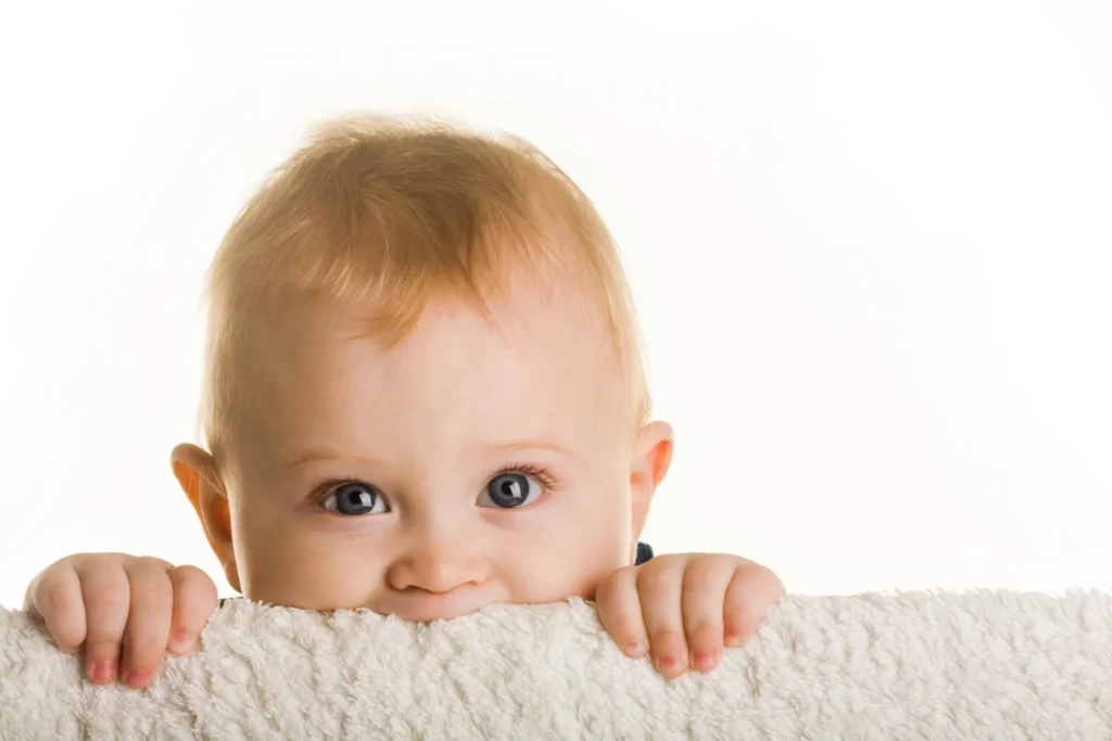 Blonde infant with blue eyes peeking over white blanket edge against white background.